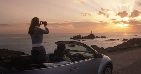 Une femme debout dans une voiture décapotable prend une photo d'un coucher de soleil sur la mer avec un phare au loin